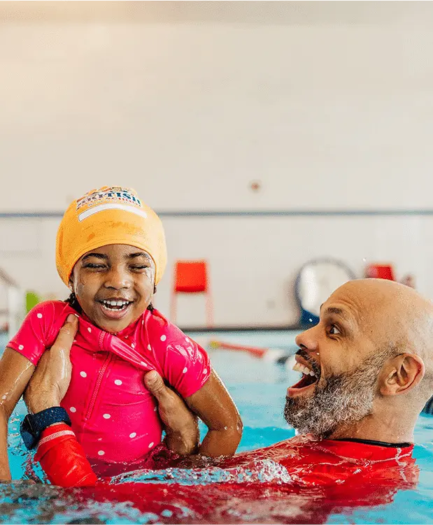 A happy young girl in a yellow swim cap is held by a smiling instructor in a