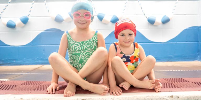 Two little girls sitting along edge of pool with swim caps and goggles and smiling