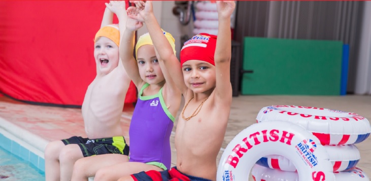Three kids sitting on edge of pool raising hands in air and smiling next to British Swim School floats