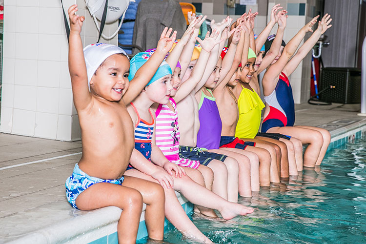 Children sitting on edge of pool holding hands in the air during swim lesson