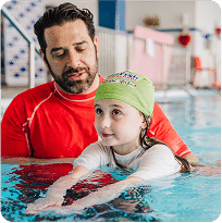 A male swim instructor helps a young girl in a green cap learn to swim in a pool.