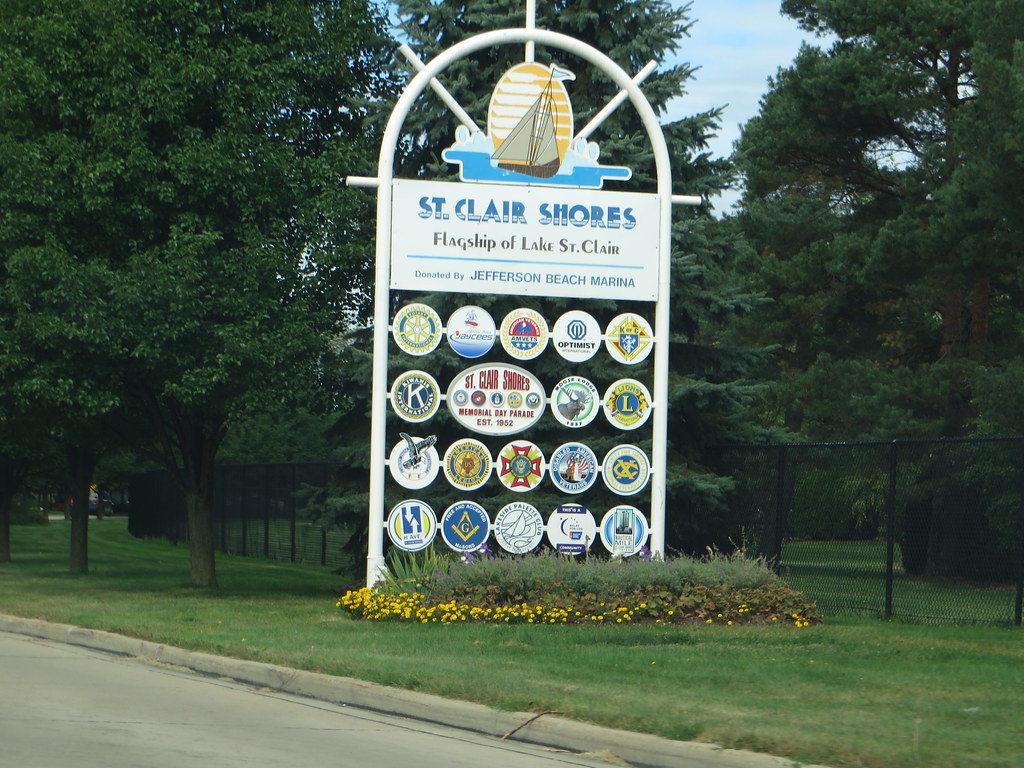 Un cartel de bienvenida en la carretera para St. Clair Shores, Michigan, con un logotipo de velero y el eslogan «Flagship of Lake St. Clair». Debajo del texto principal, varios emblemas circulares representan a varias organizaciones comunitarias y de servicios locales.