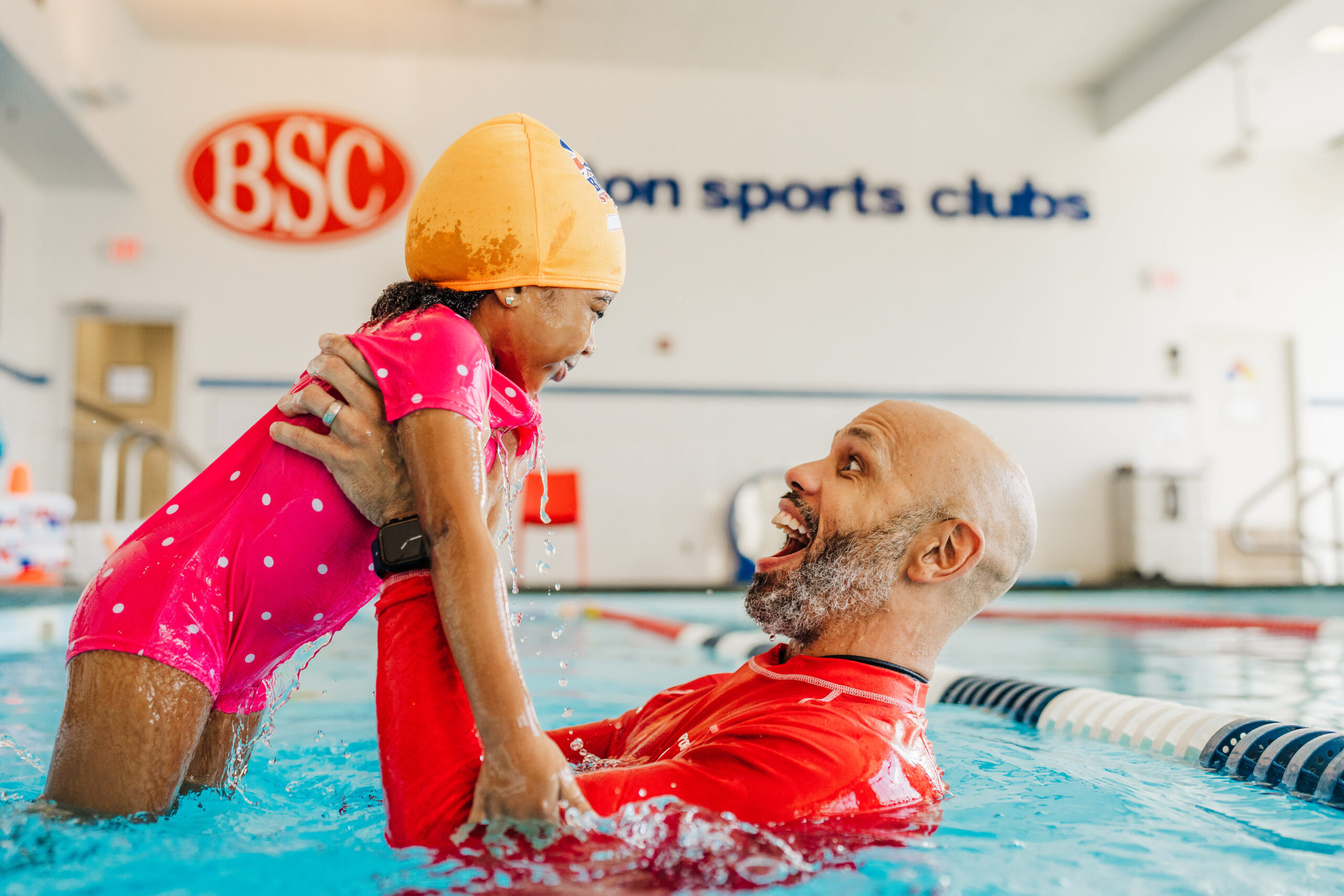 A happy instructor holds a smiling child in a bright indoor swimming pool.