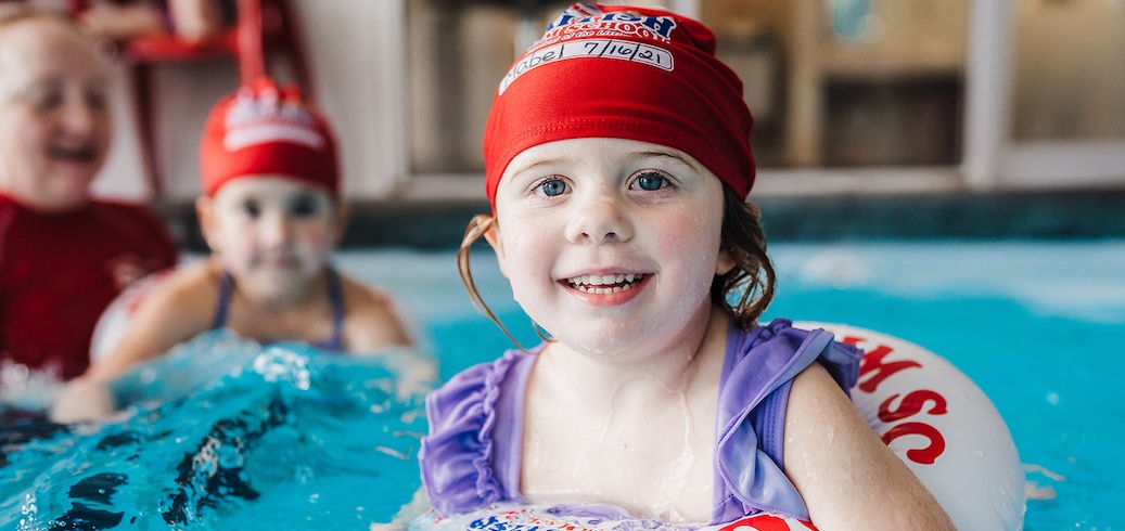 A cheerful girl with a red swim cap and purple swimsuit smiles in a blue swimming pool.