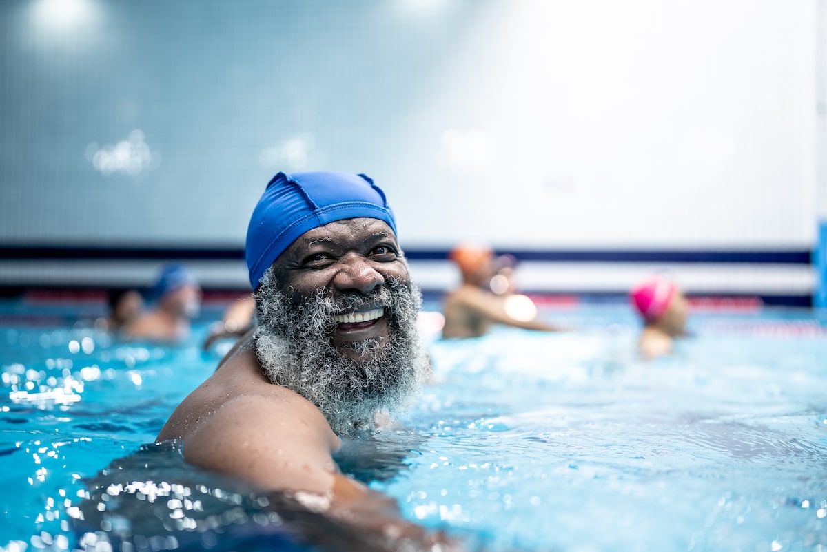 A smiling senior Black man with a white beard and blue swim cap enjoys a group swim.