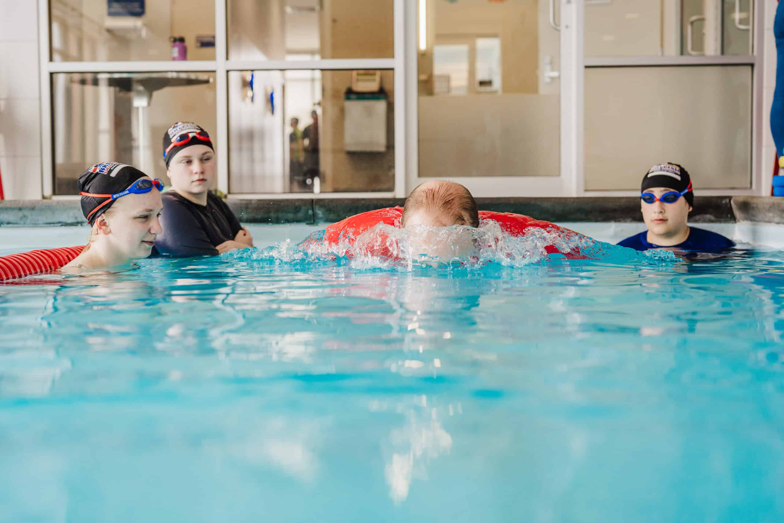 Three children in swim caps and goggles practice in a pool, one wearing a red learn-to-swim float.
