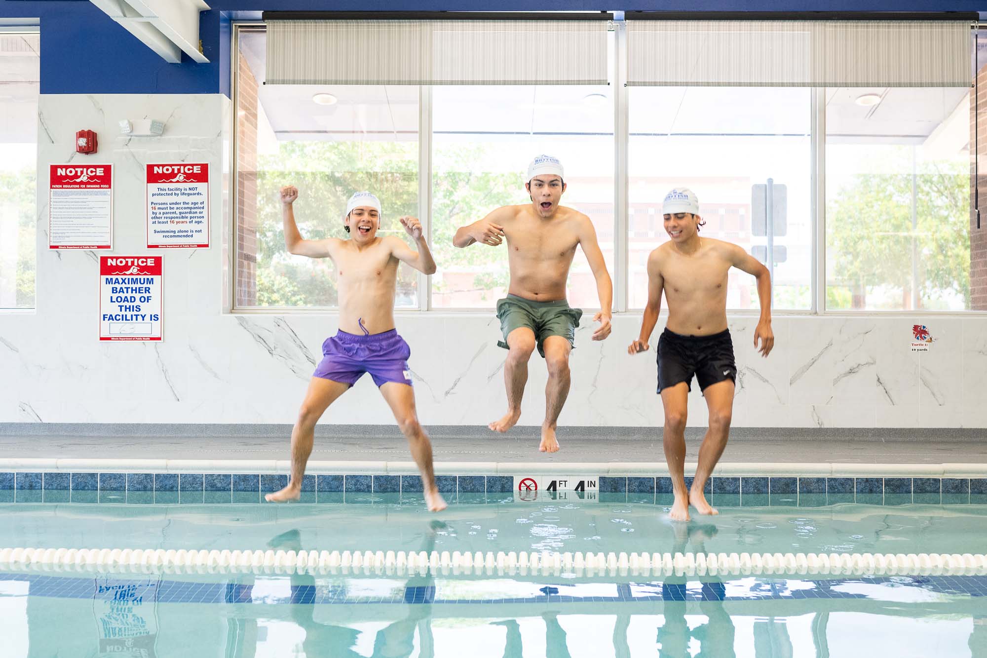 Three young men in swim caps jump joyfully into an indoor swimming pool. TITLE