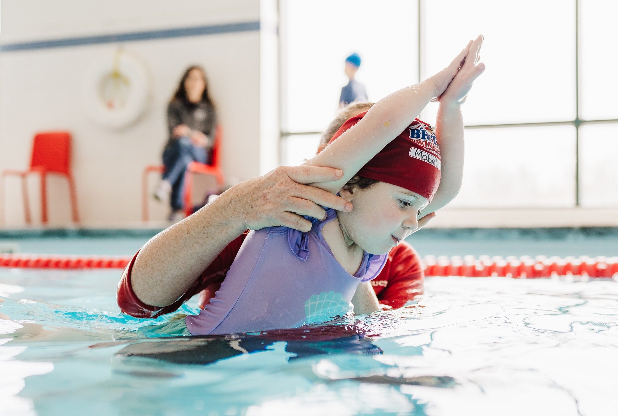 A child in a red swim cap practices swimming strokes with an instructor's help