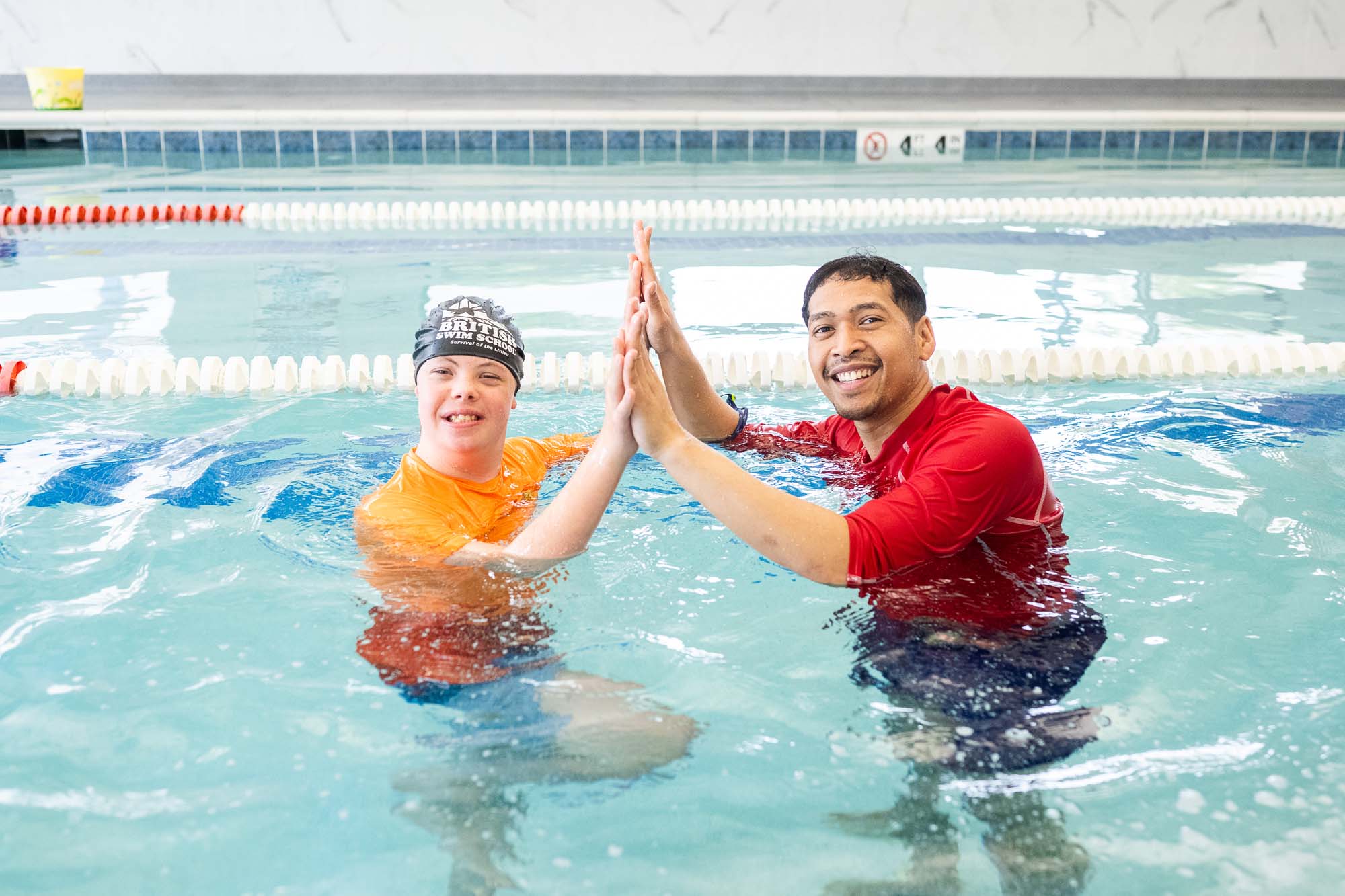 A smiling young swimmer and an adult instructor high-five in a bright indoor pool. TITLE