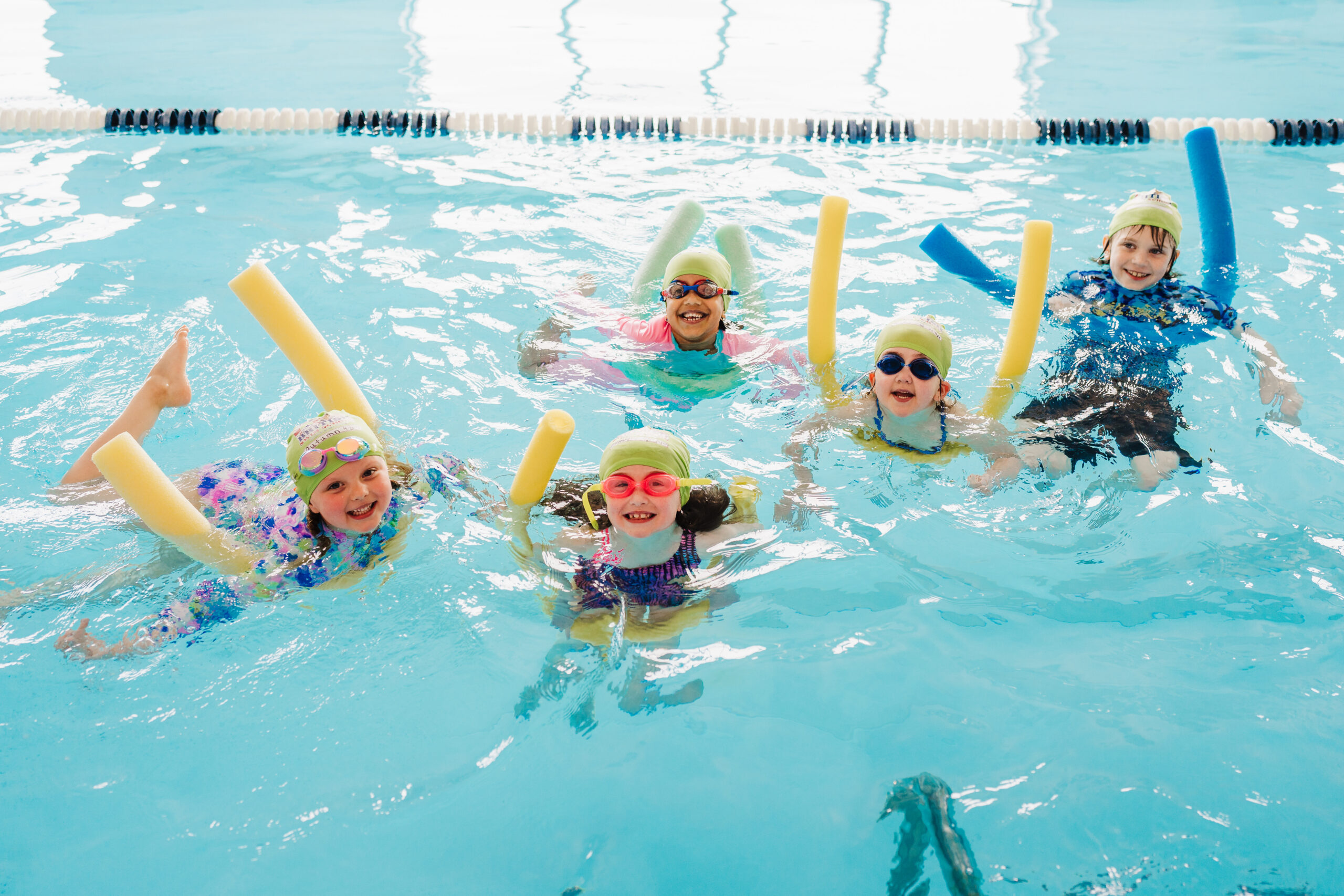 A group of five smiling children in swim caps and goggles holding pool noodles during a British Swim School group swim lesson in an indoor pool.