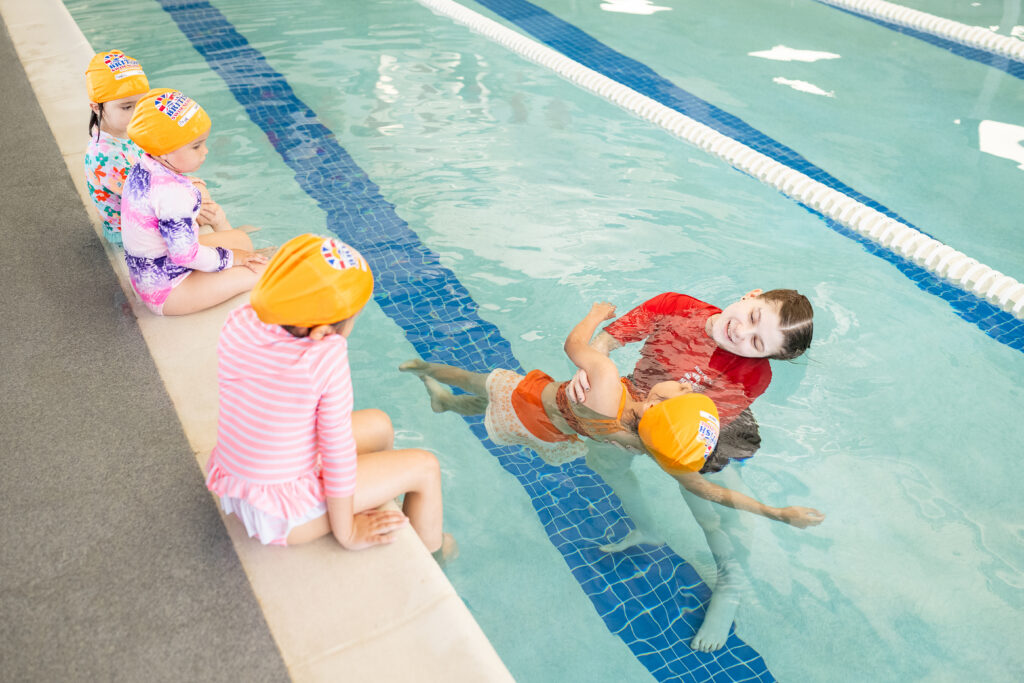 A British Swim School instructor gently guiding a young child through a back float while three toddlers in swim caps wait at the pool's edge.