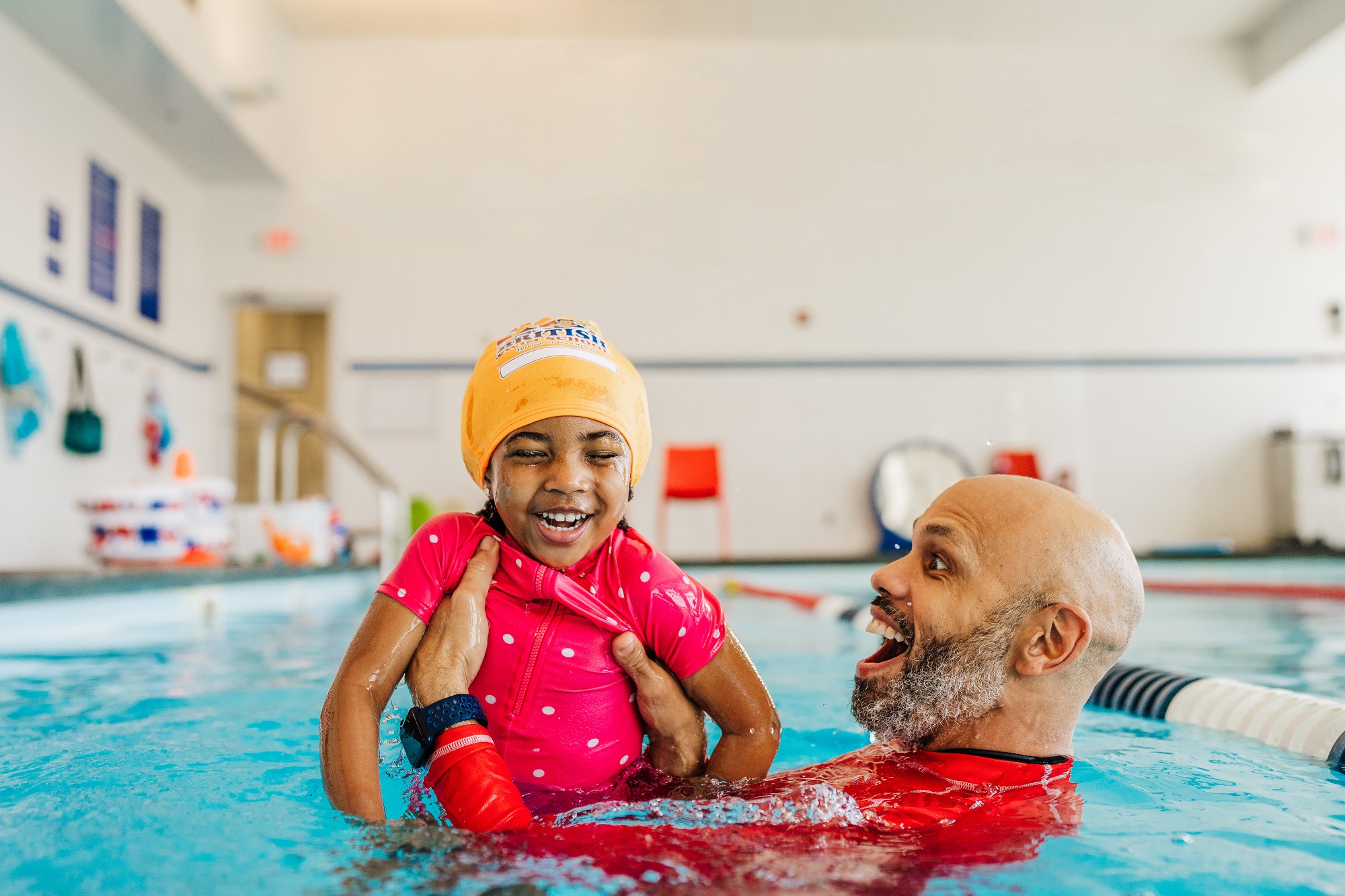 A joyful child in a pink swimsuit and yellow cap laughs with an adult in a swimming pool.