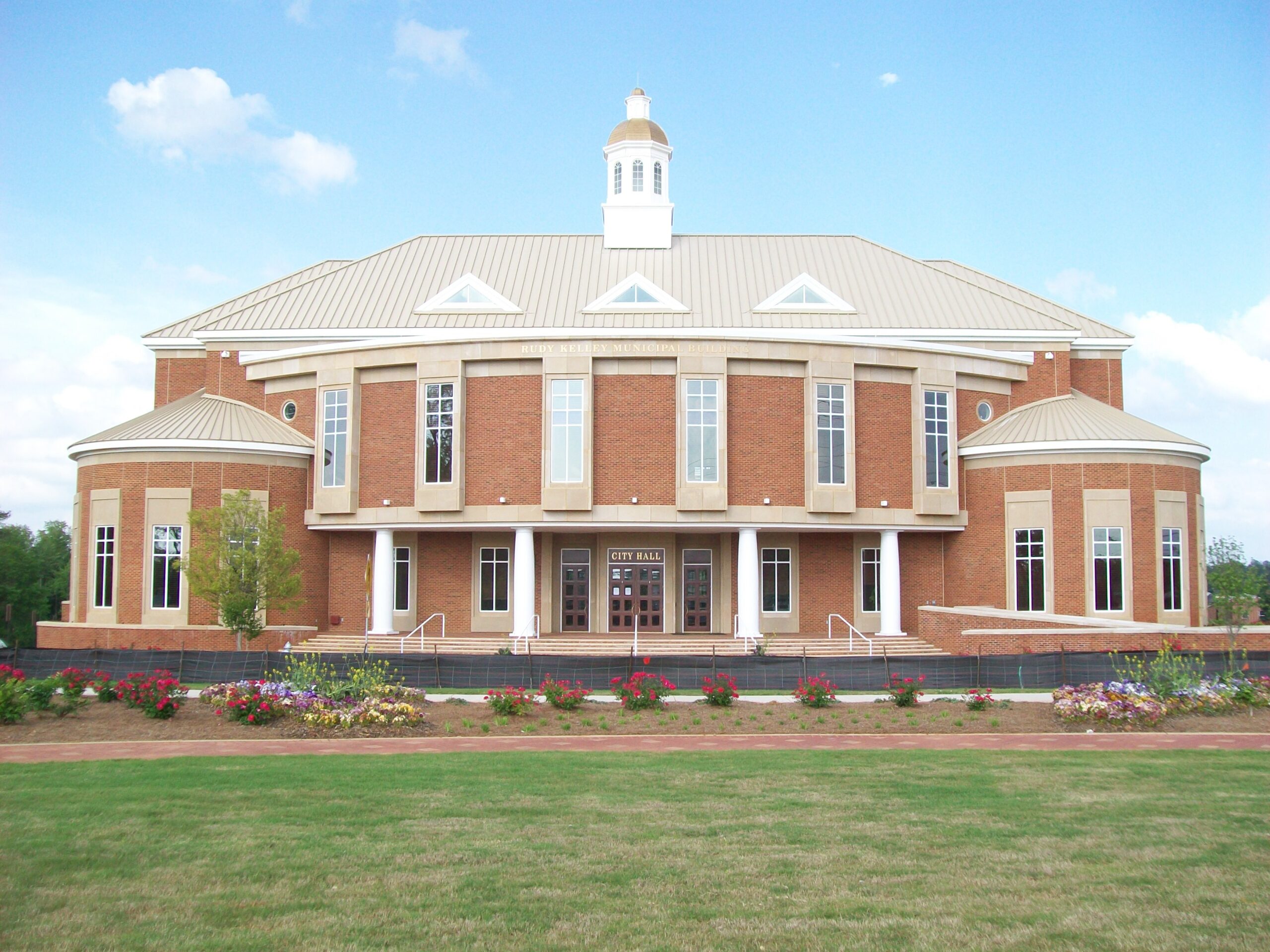 A modern brick city hall with a central cupola, tan roof, and vibrant flower beds in