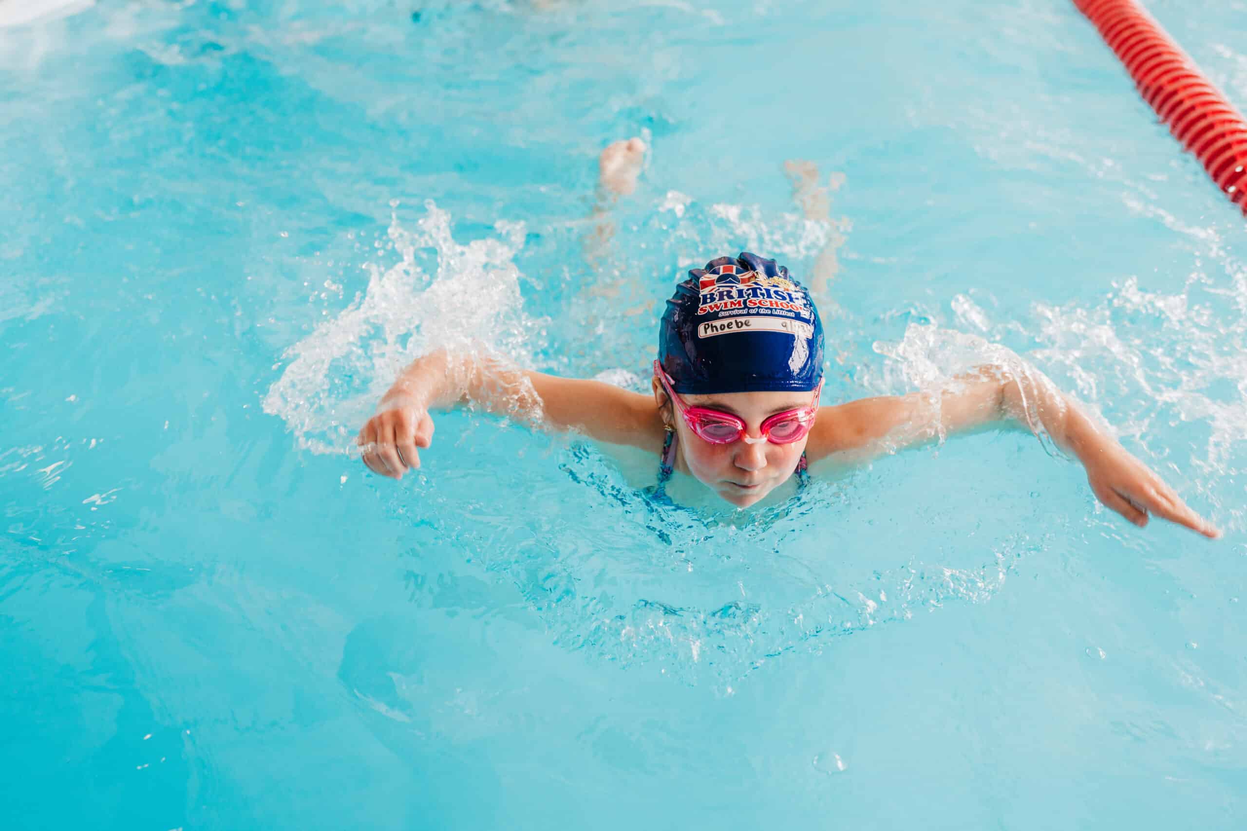 A young swimmer in a blue cap and pink goggles swims butterfly in a bright blue pool.