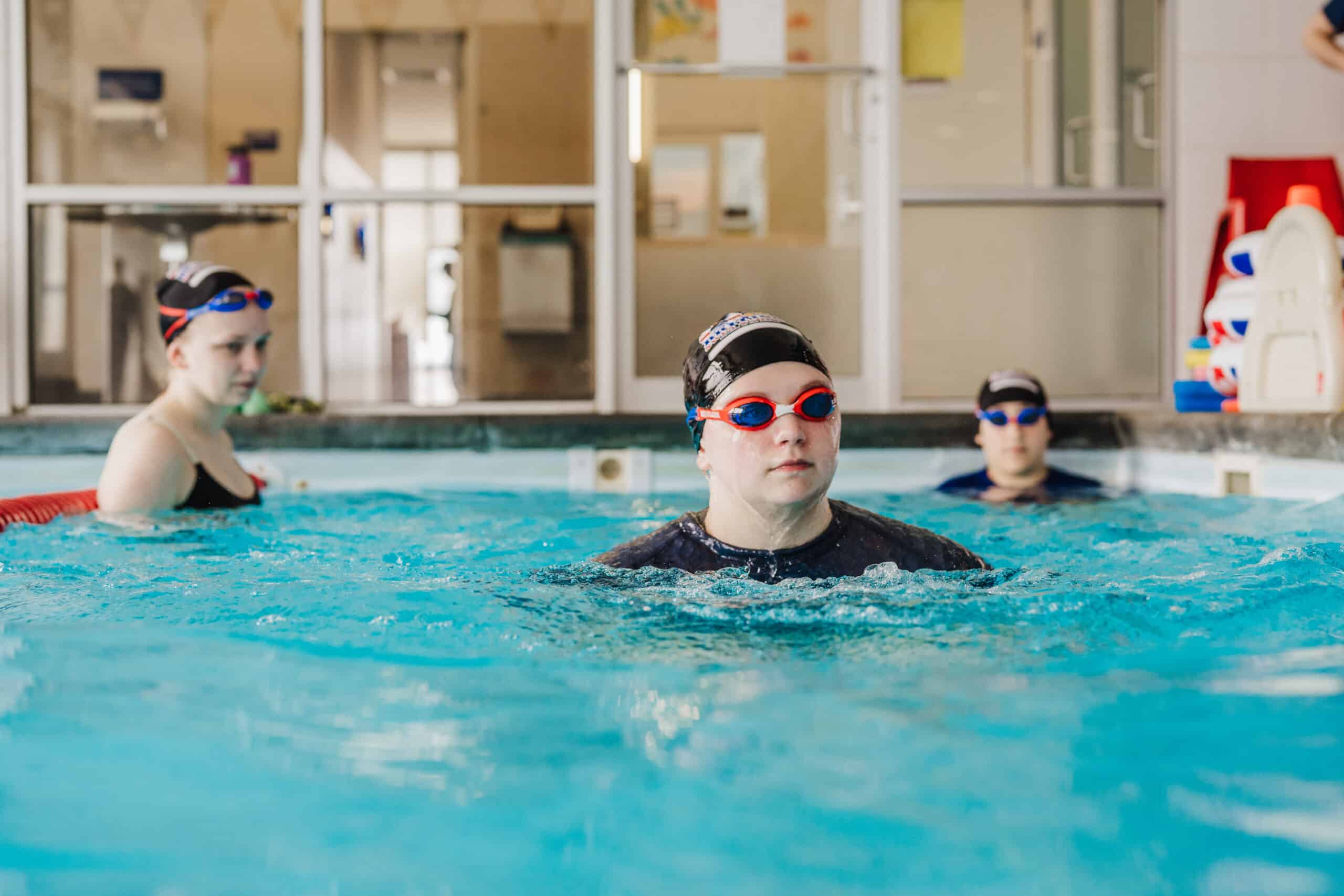 Three young swimmers in caps and goggles stand in a bright indoor pool.