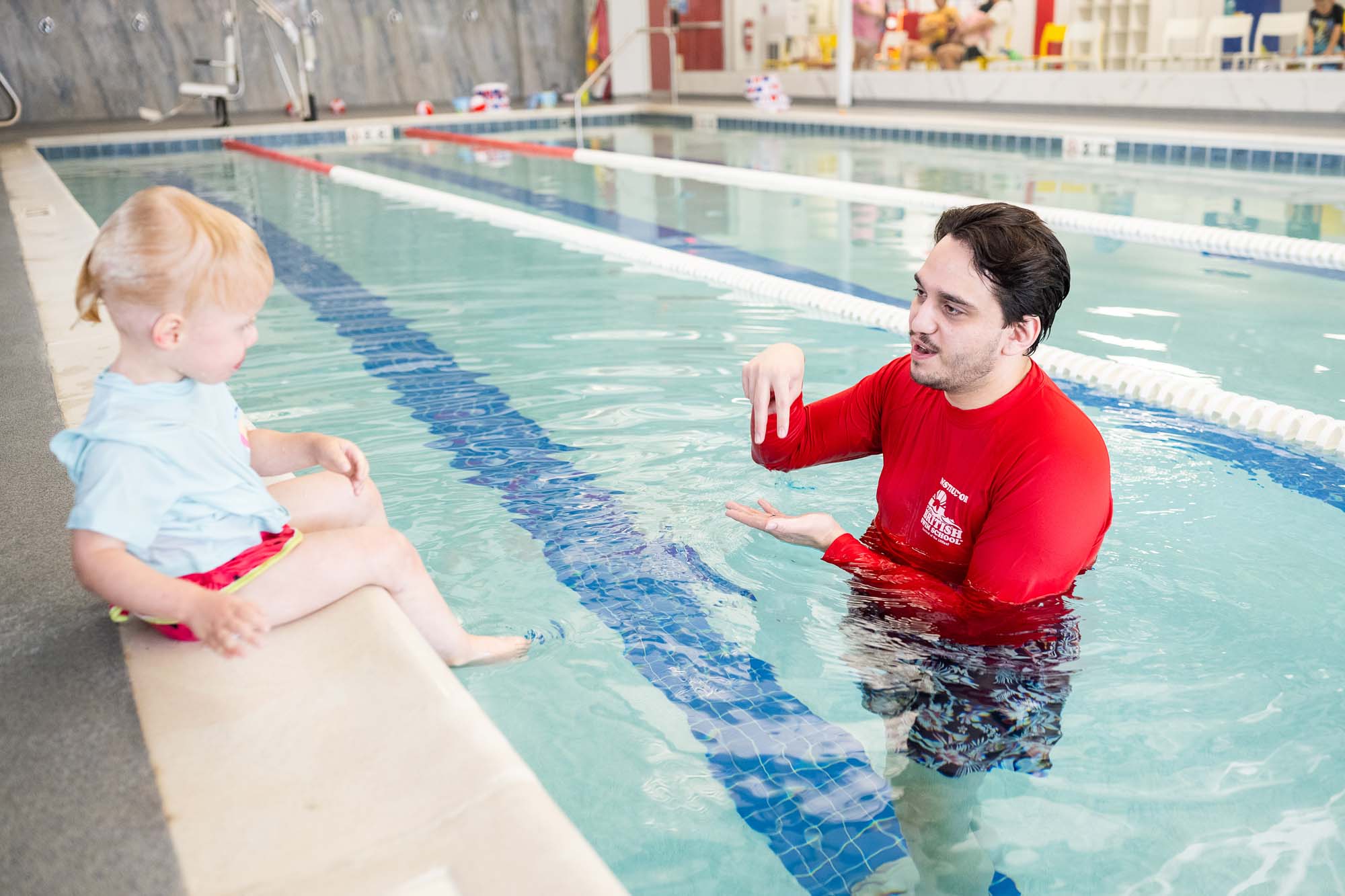 A swim instructor teaches a young child sitting on the edge of an indoor pool. TITLE: