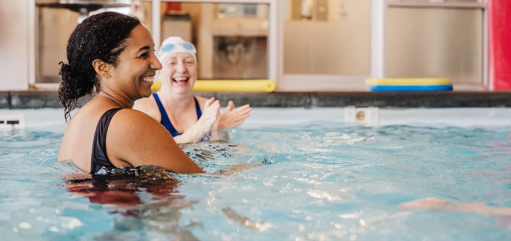 Two smiling women enjoy a swim in a pool, one laughing and clapping.