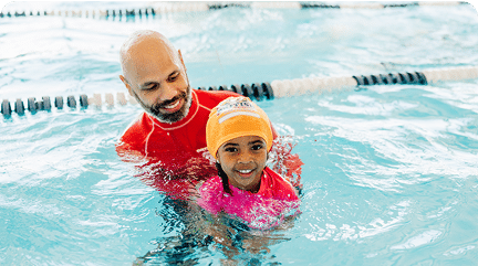 A smiling swim instructor supports a young girl wearing an orange cap in a pool. TITLE: