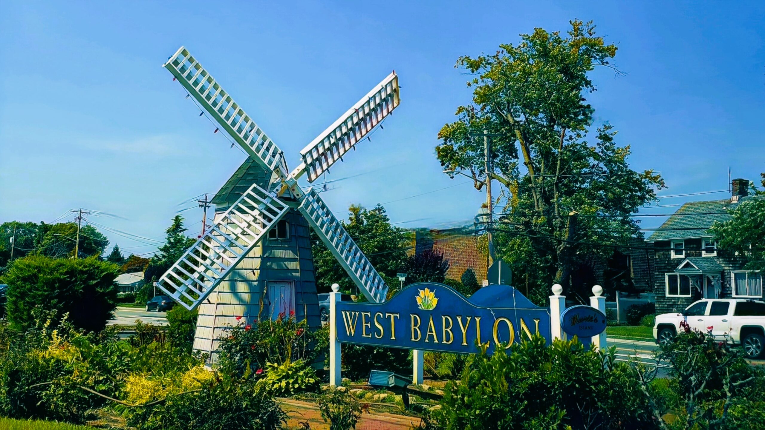 A charming blue windmill and "WEST BABYLON" sign are surrounded by greenery under