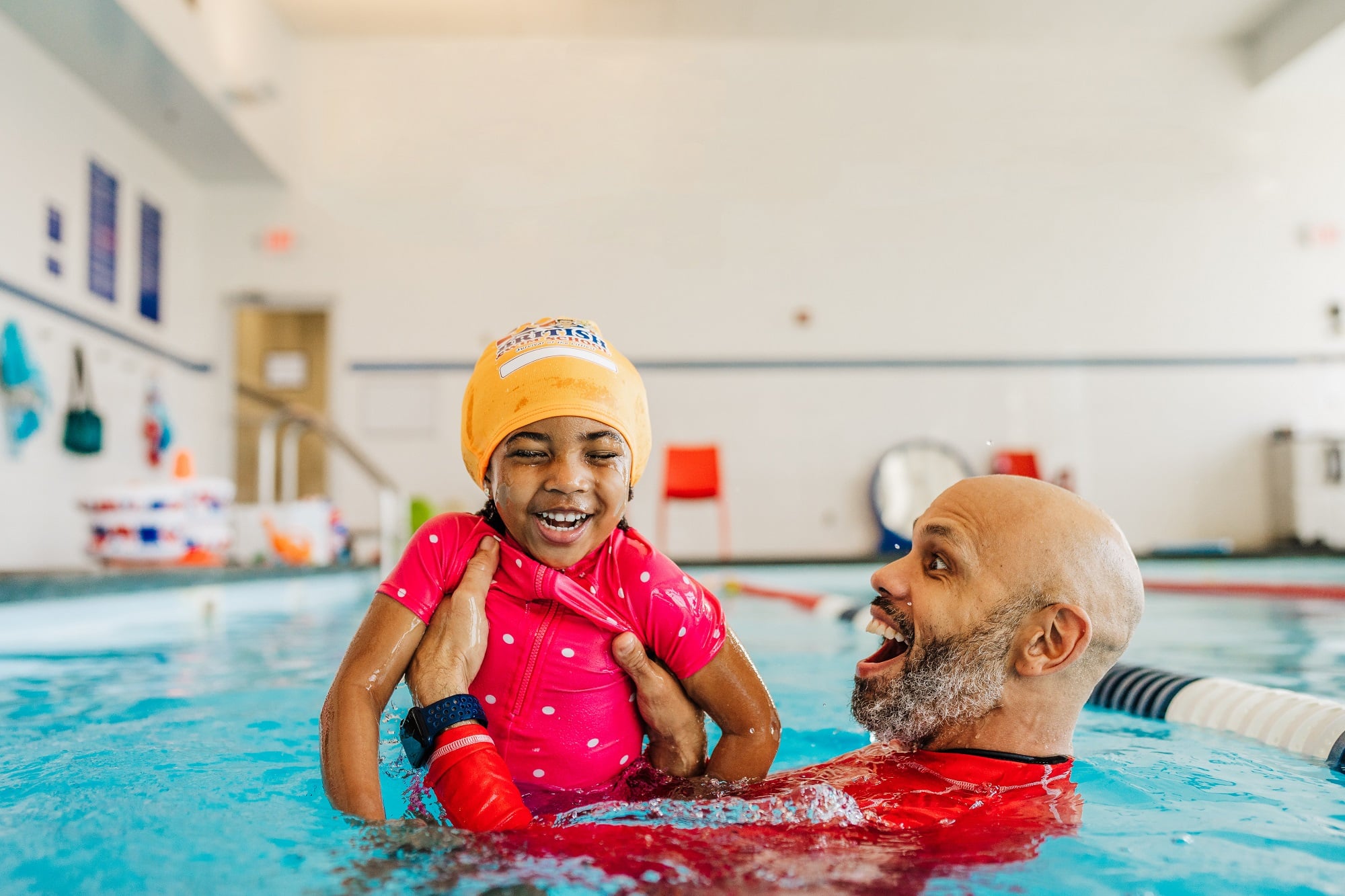 A joyful child in a pink swimsuit and yellow cap laughs with an adult in a swimming pool.