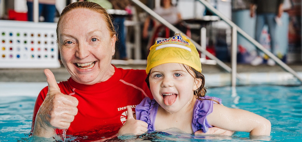 A smiling swim instructor and young child give thumbs-up in a swimming pool. TITLE: