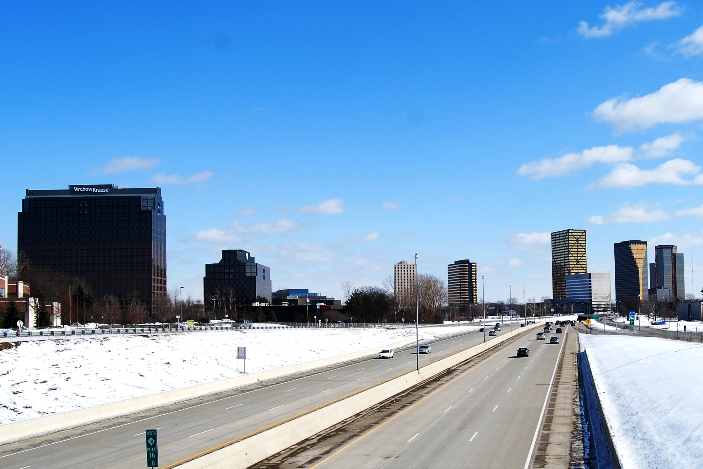 A wide highway with cars and snow-covered shoulders leads towards a city skyline under a bright blue