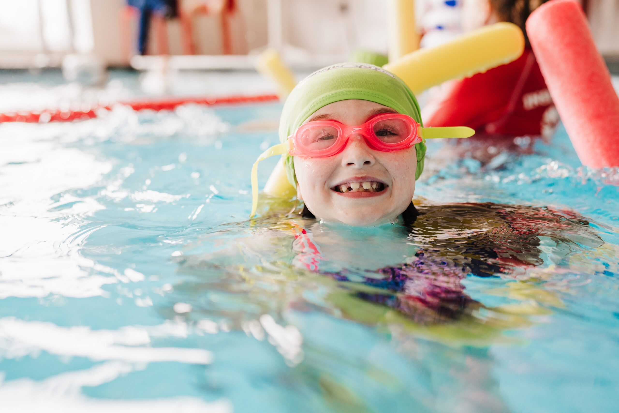 A happy child in a green swim cap and pink goggles smiles in a swimming pool.