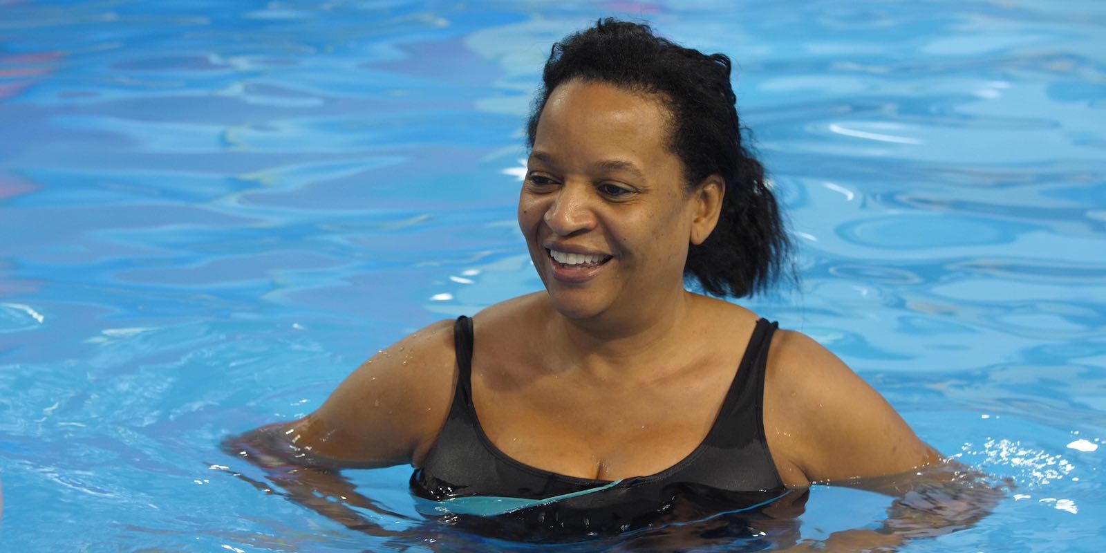 A smiling Black woman in a black swimsuit stands happily in a bright blue swimming pool.