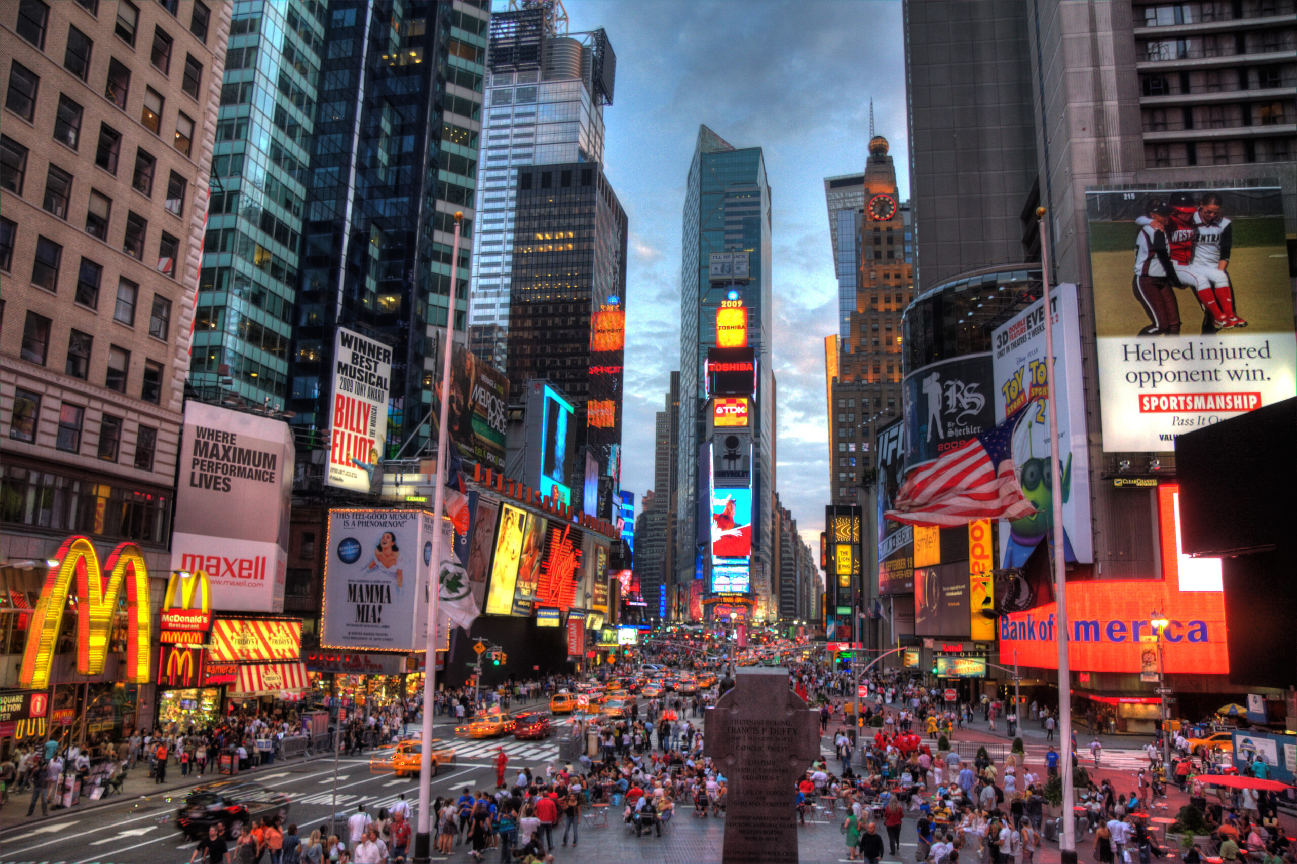 Times Square at dusk, filled with bright digital billboards, bustling crowds, and yellow taxis