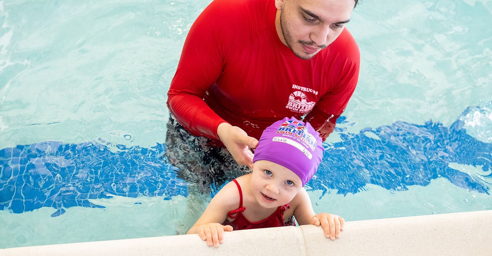 A swim instructor in a red shirt helps a young girl in a purple British Swim School cap as she holds onto the edge of the pool.