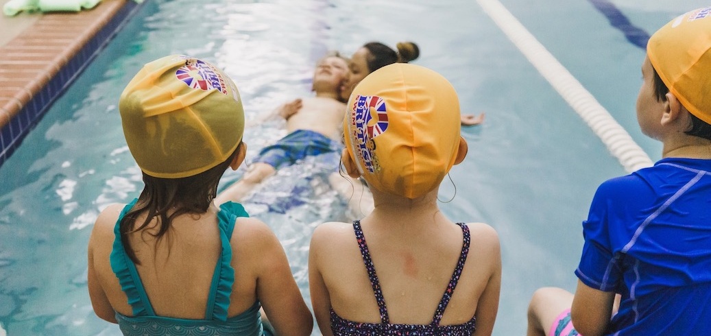 Three children in swim caps sit poolside, watching two people float on their backs in the water