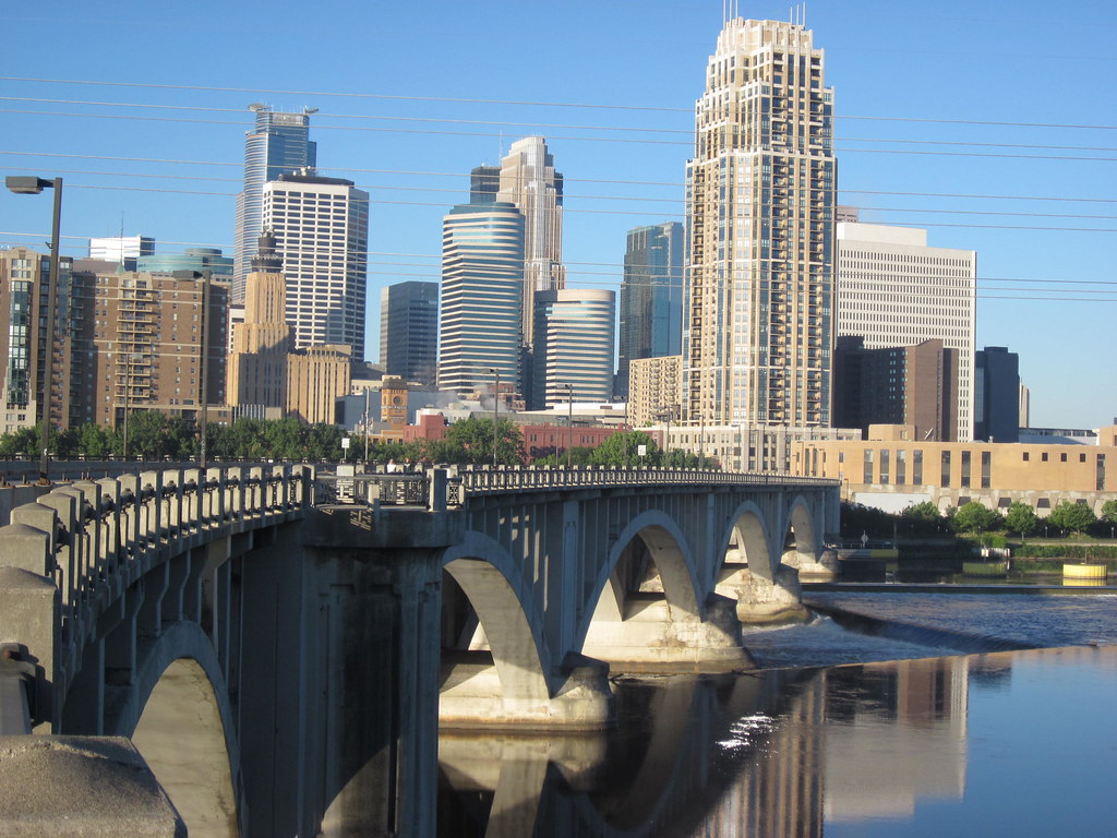 The iconic Stone Arch Bridge crosses the Mississippi River with the Minneapolis skyline under a clear blue sky.