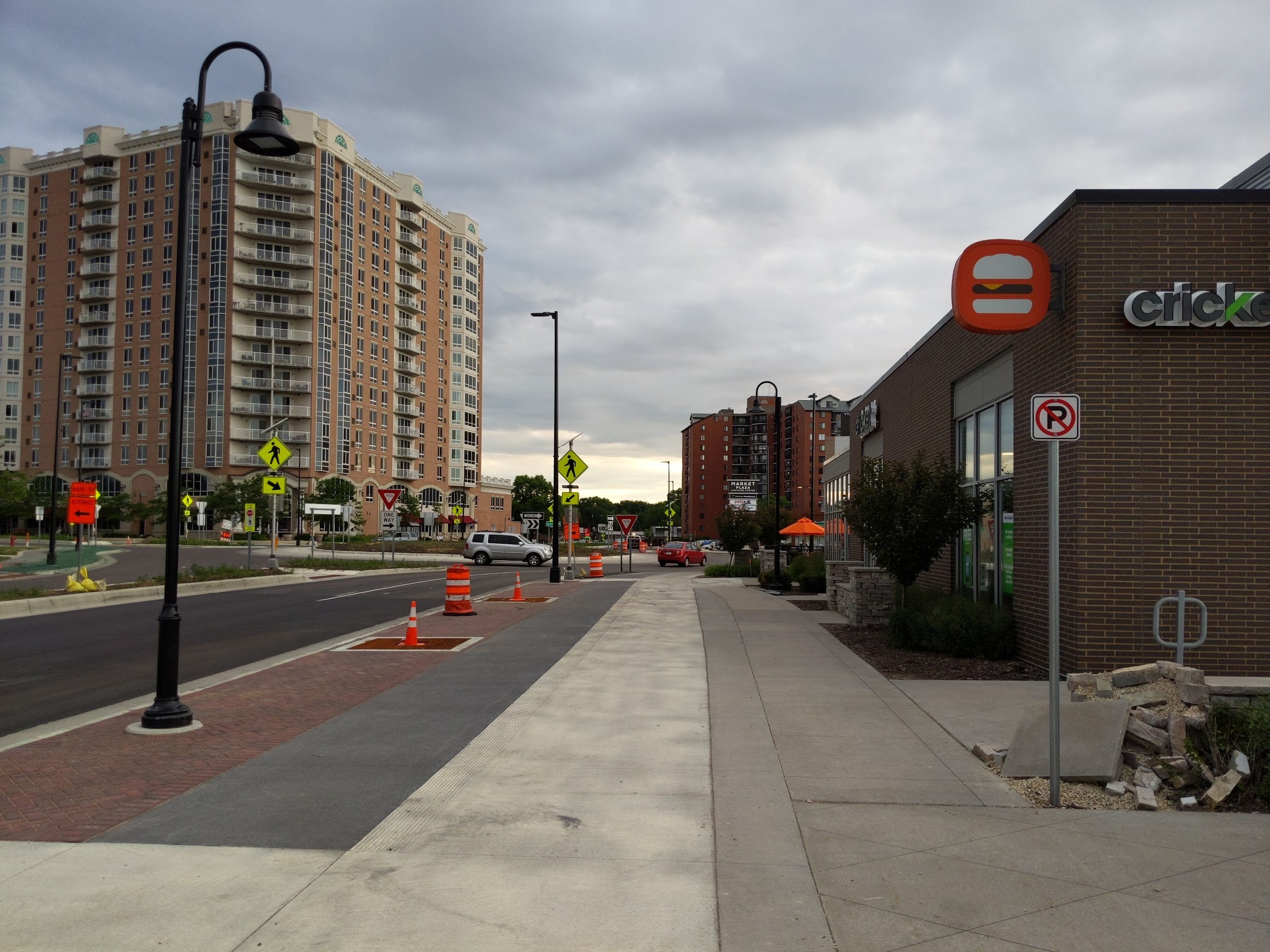 A cloudy urban street features a tall apartment building, a commercial strip with a burger sign,