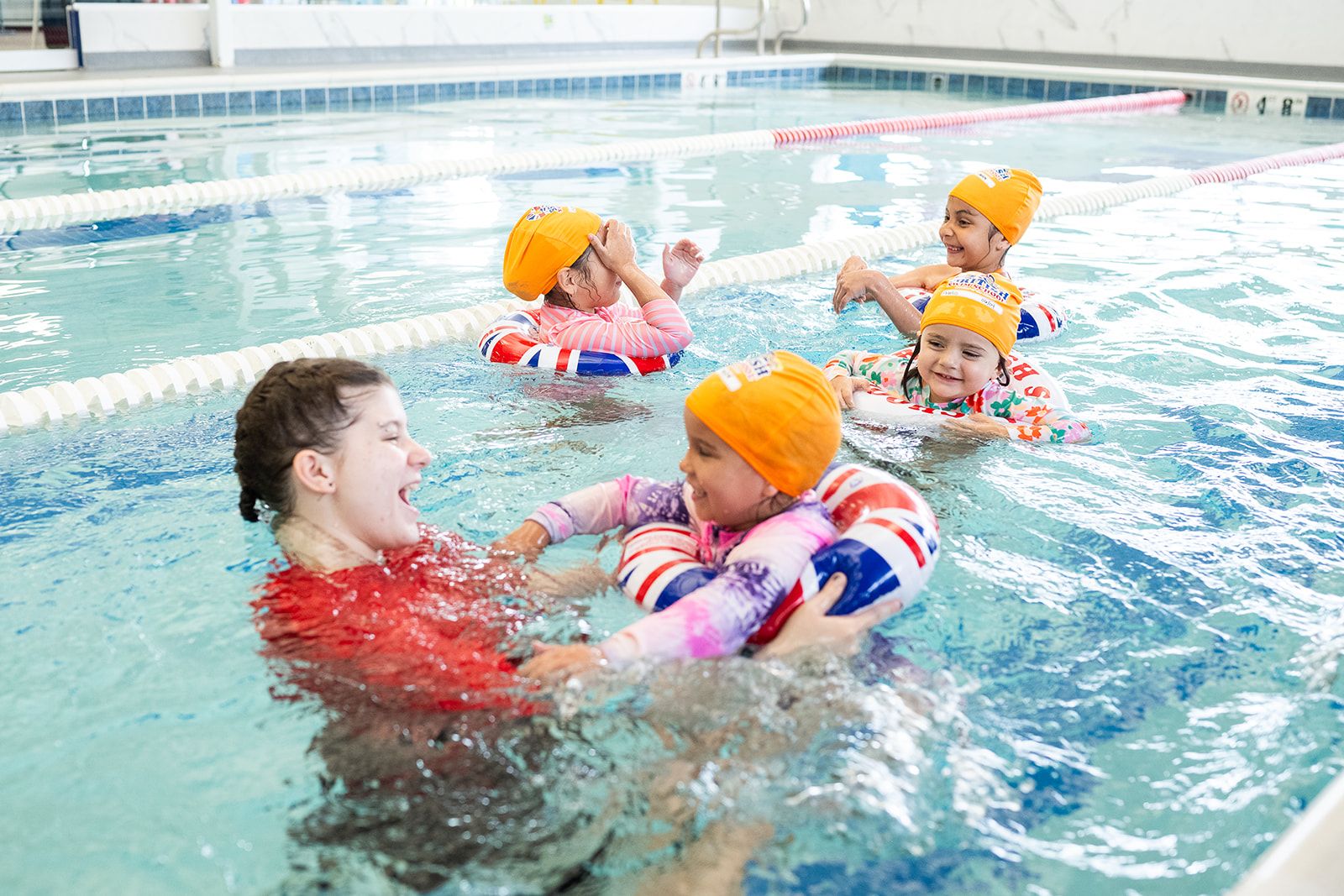 A British Swim School instructor laughs while helping a young girl during a kids group swim lesson.