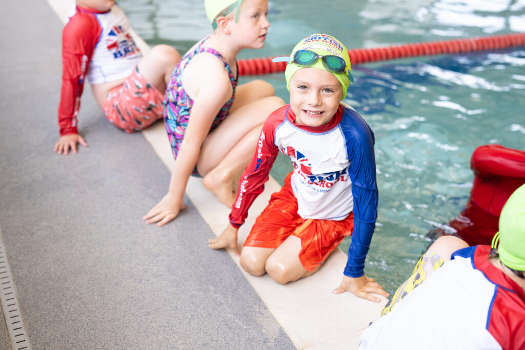 A smiling young boy in a branded British Swim School rash guard and green swim cap sitting on the edge of an indoor pool during a group swimming lesson with other children.