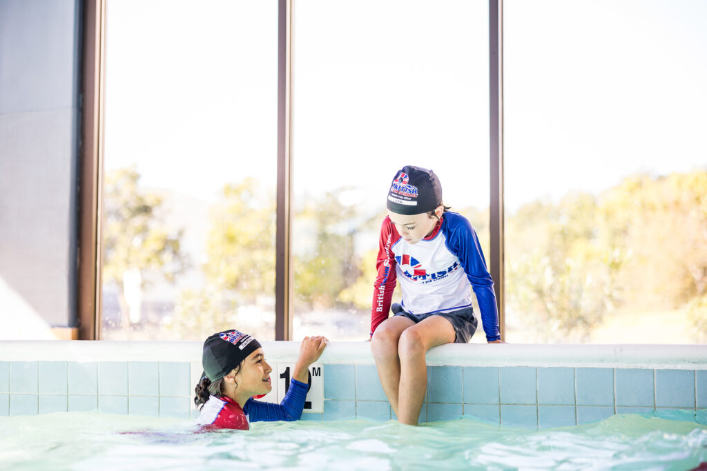 Two children participating in British Swim School swim lessons in Montgomery County, PA, with one child in the indoor pool and another sitting on the edge wearing matching swim caps and rash guards.