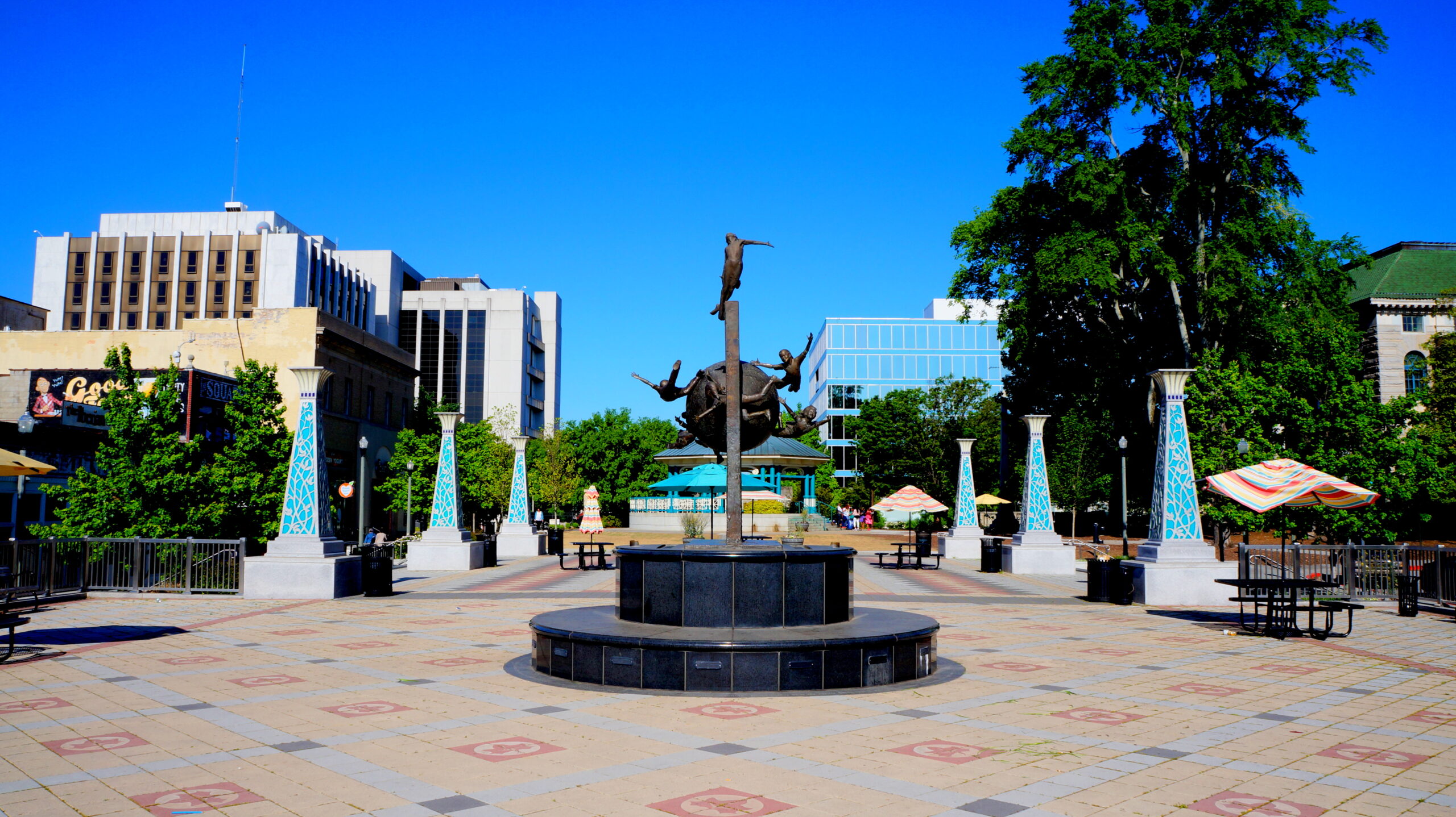 A large bronze sculpture of figures on a sphere dominates a sunny city plaza with buildings and trees.