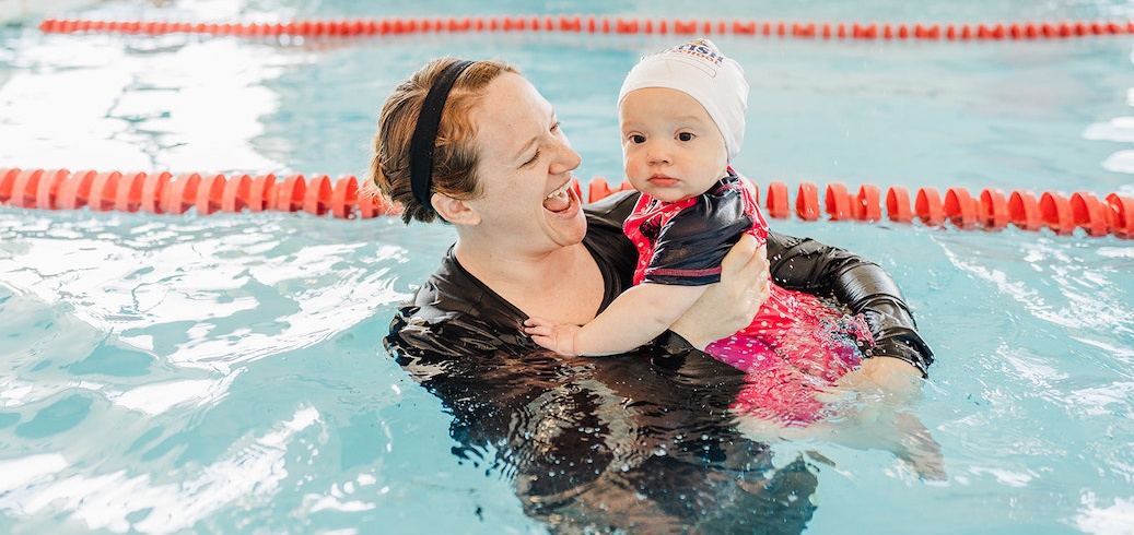 A joyful woman holds a baby in a swim cap during a swimming lesson. TITLE: