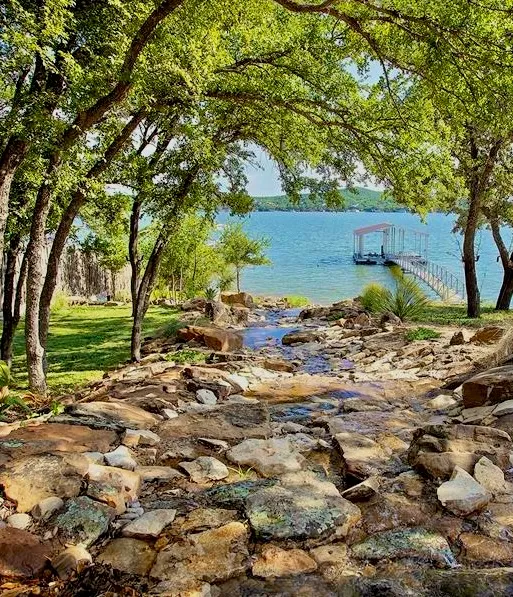 Lush green trees frame a rocky stream leading to a serene lake with a boat dock.