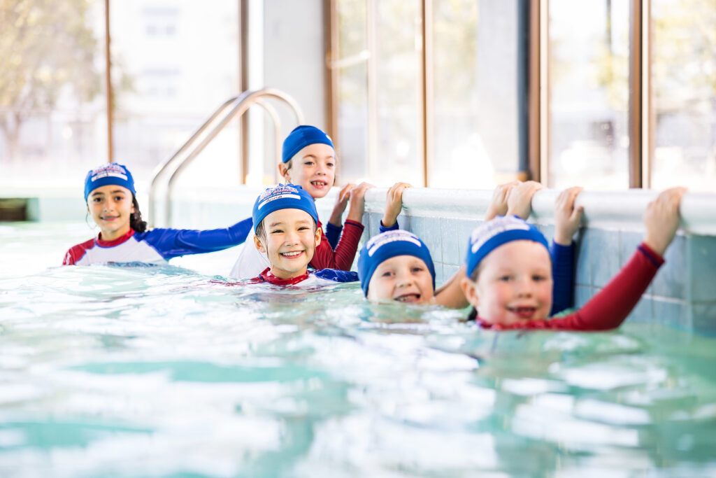 Five children in British Swim School caps and rash guards hold the pool wall and smile during a small-group lesson at an indoor pool.