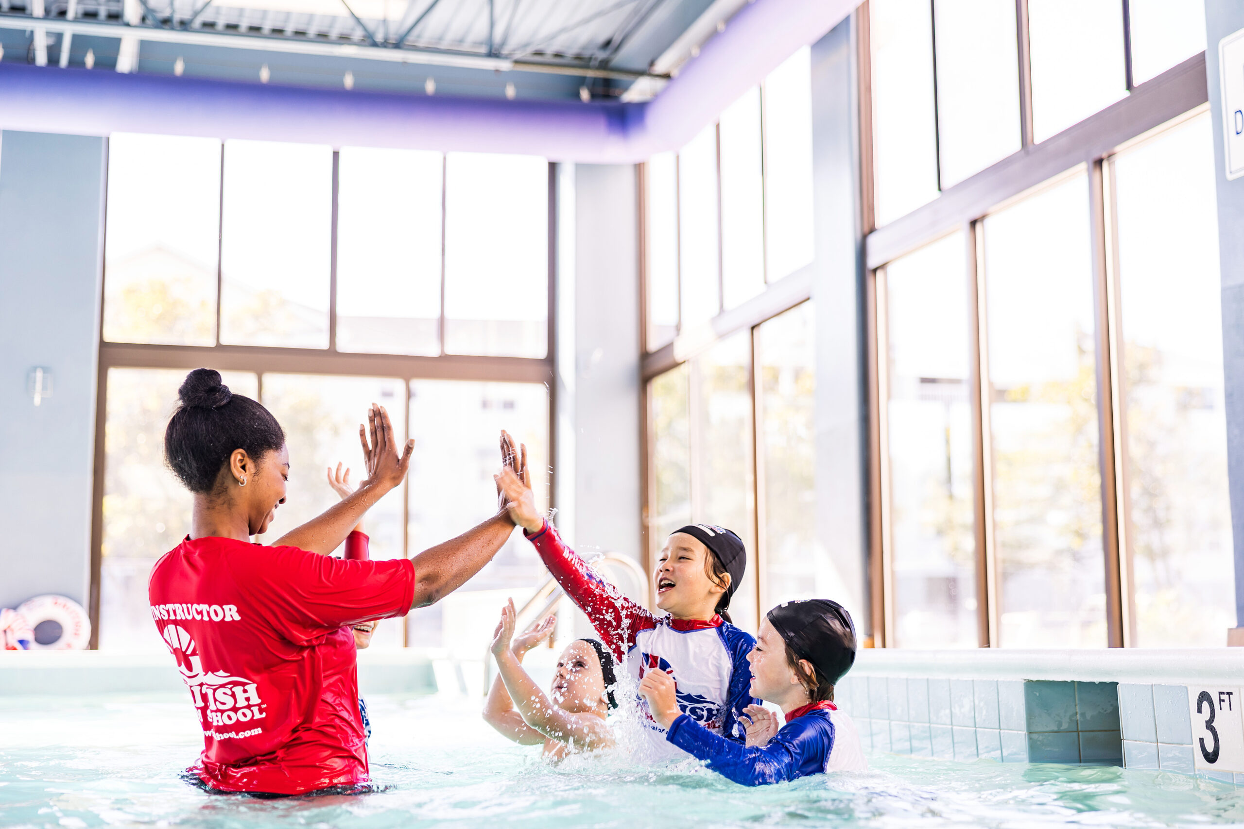 A British Swim School instructor high-fives a small group of smiling children during a swim lesson in an indoor pool.