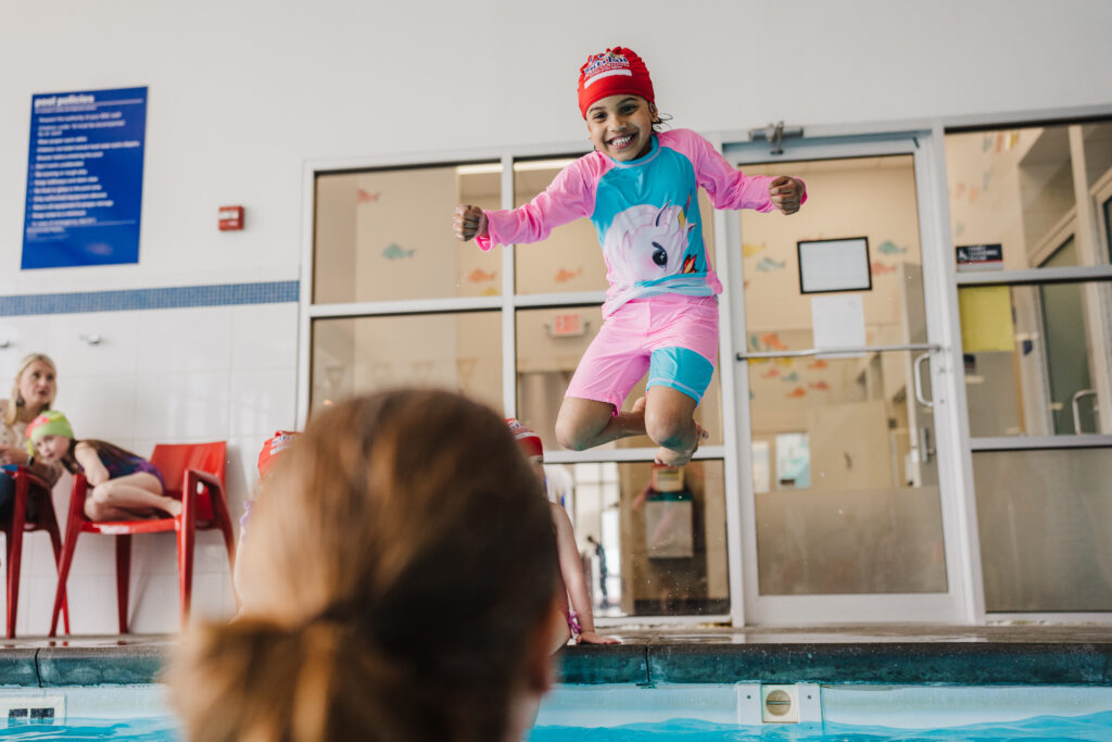 A grinning girl in a unicorn rash guard and red cap jumps toward the water and her instructor at an indoor British Swim School pool.