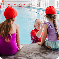 A smiling swim instructor high-fives a young girl sitting by the pool while another watches.