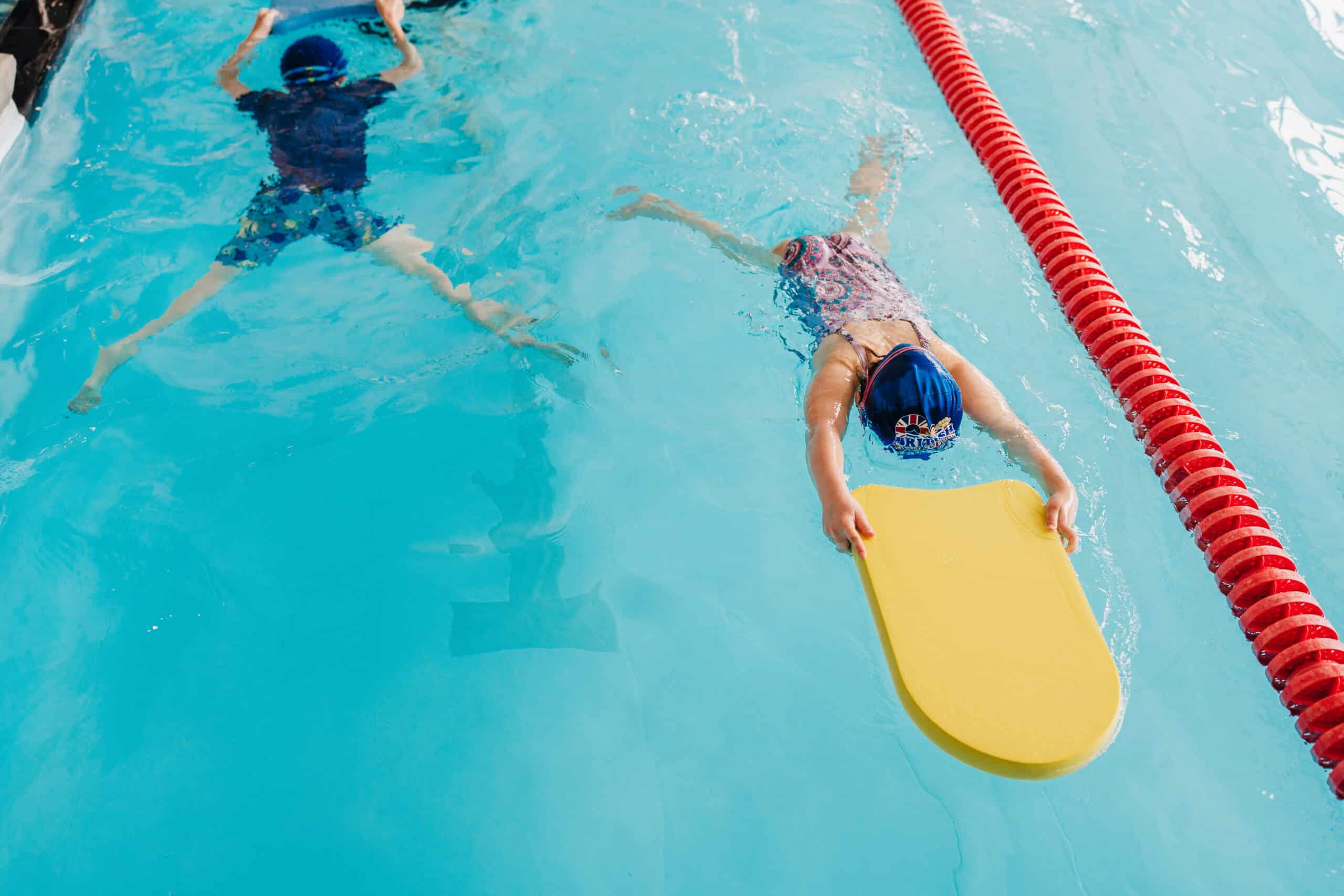 A child in a blue cap uses a yellow kickboard to swim in a pool, with another