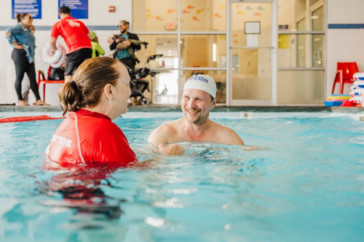 A smiling swim instructor talks to a happy man in a swim cap in an indoor pool.