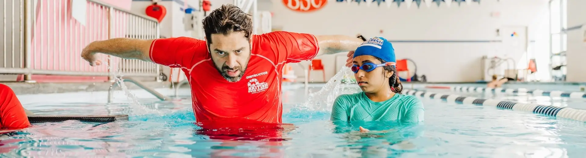 A male swim instructor in a red shirt demonstrates a stroke to an attentive female student in a pool