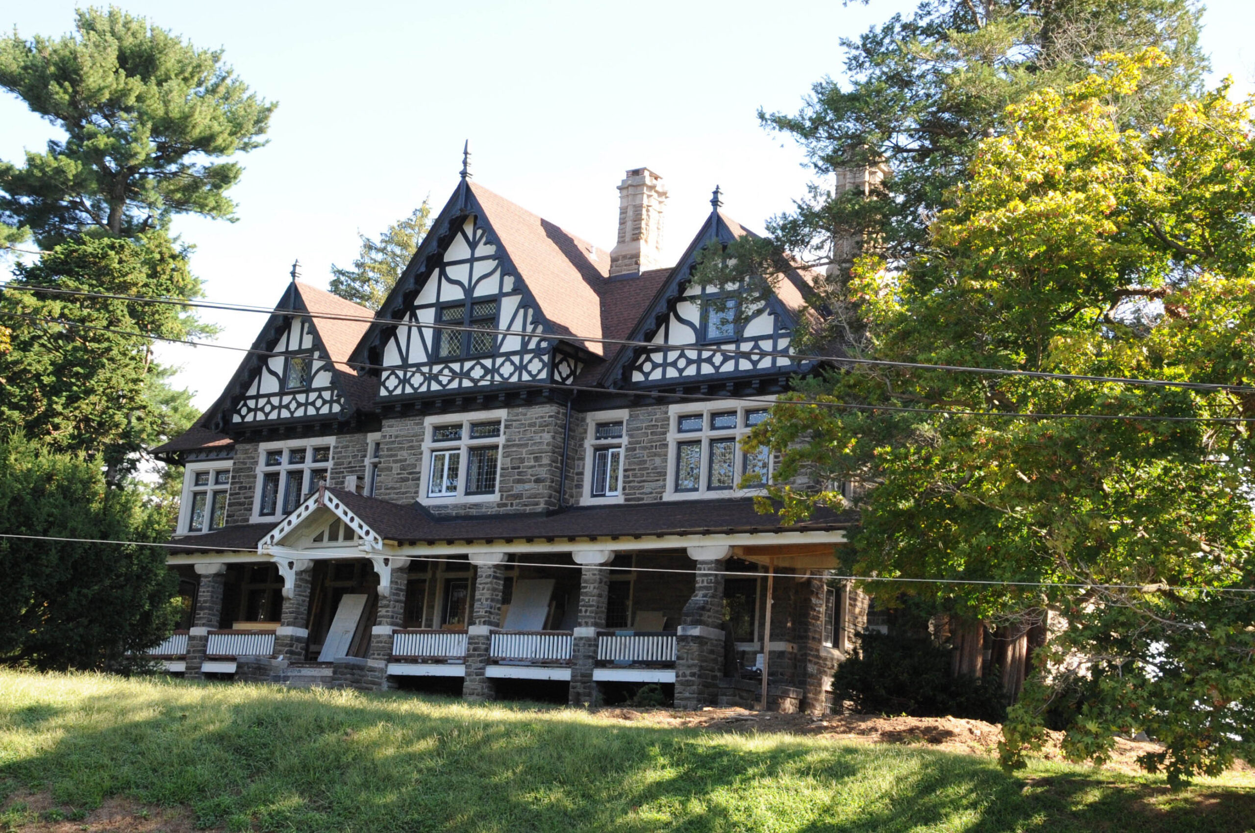 The exterior of Fairlawn Manor in Philadelphia, a historic Tudor Revival mansion featuring a Wissahickon schist stone facade, intricate half-timbered gables, a wide covered front porch, and tall decorative chimneys surrounded by mature trees.