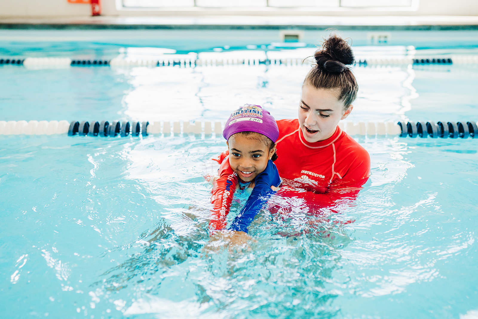 An instructor helps a smiling child learn to swim in a bright indoor pool. TITLE