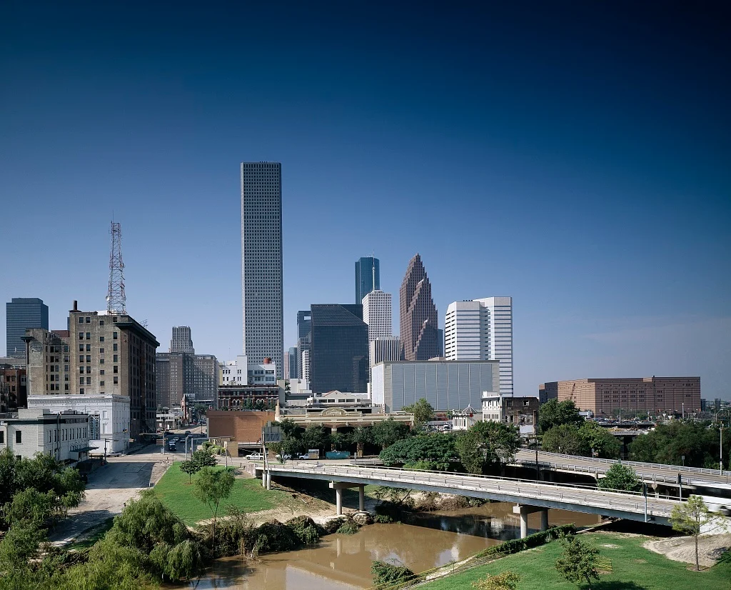 A panoramic view of the Downtown Houston skyline, featuring landmarks like the JPMorgan Chase Tower and Wells Fargo Plaza, as seen across the Buffalo Bayou with its bridges and the historic near-Heights area. British Swim School offers year-round, indoor swim lessons for the Harris County community and surrounding Houston Heights, River Oaks, Midtown, and The Heights neighborhoods.