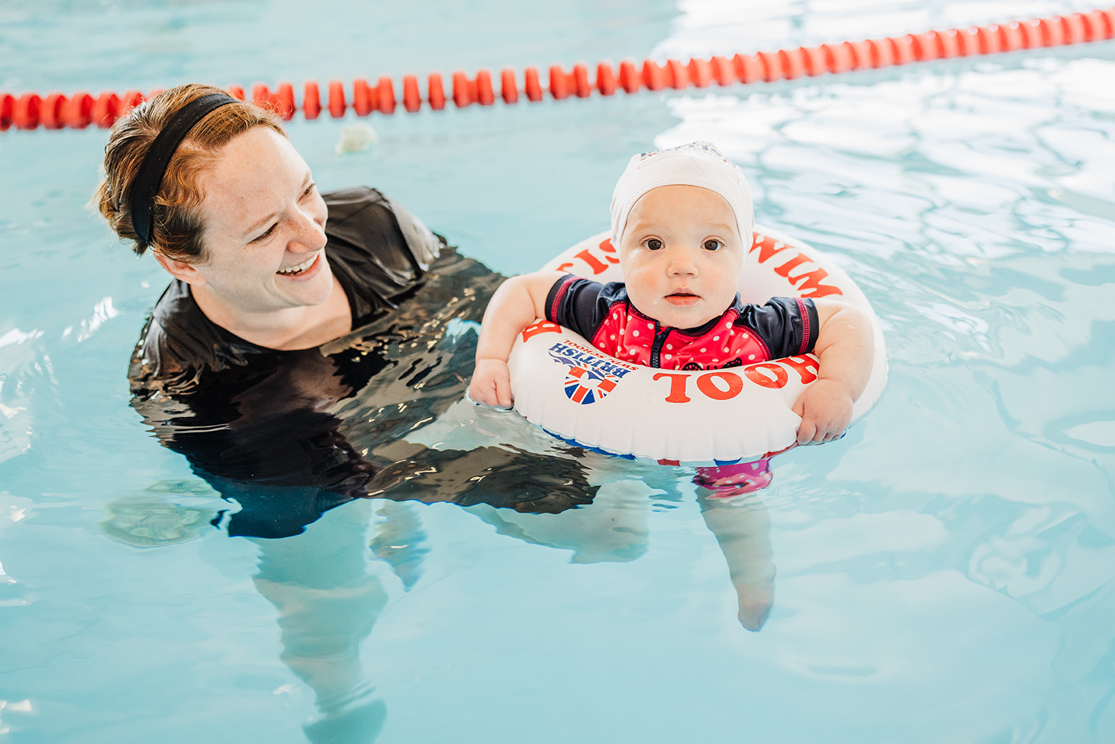 A smiling woman helps a baby in a swim ring learn to swim in a pool.